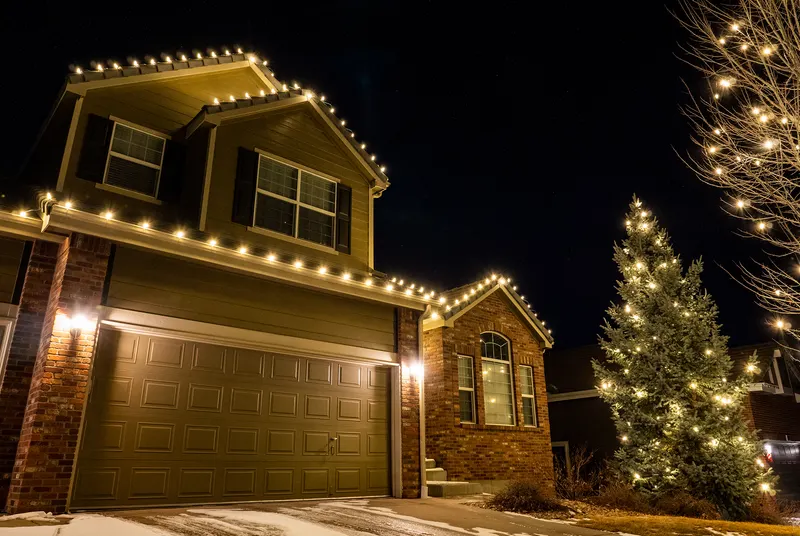 Two-story home with warm white roofline lights and a wrapped evergreen